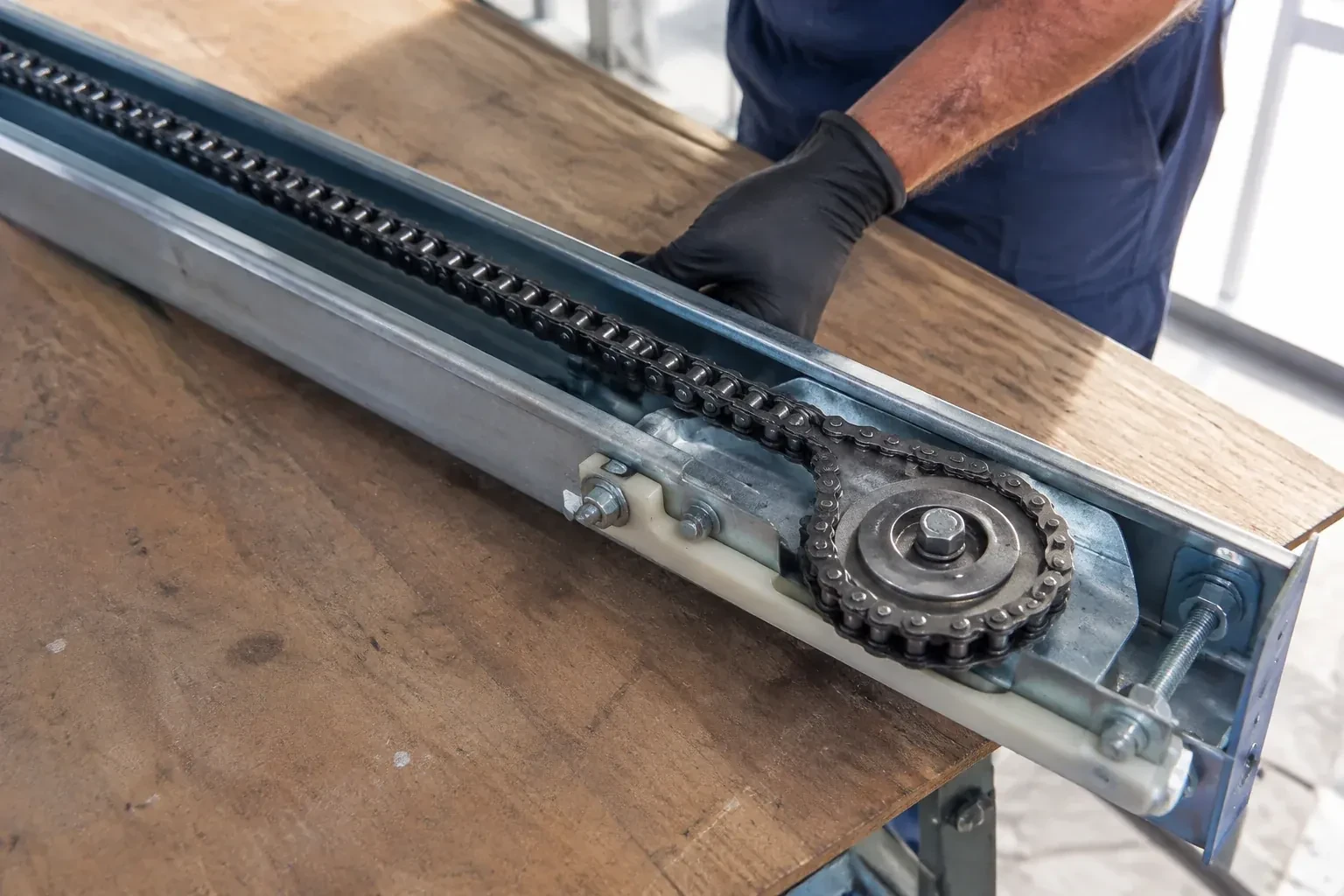 Gloved hand resting on a steel opener rail laid on a wooden bench, showing a drive chain looped around a sprocket wheel. — Chain-Drive Opener Repair, Lynden