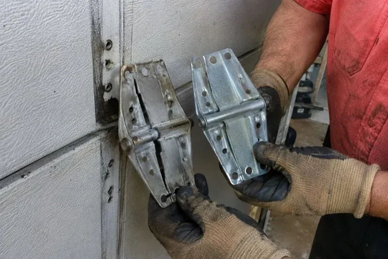 Gloved hands holding two metal garage-door hinges side by side, one discolored with a visible crack and the other bright silver. — Garage Door Hinge Replacement, Hamilton